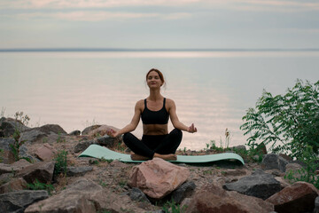 Fototapeta na wymiar Woman in black sport suit is doing meditation, yoga pose (asana), work out outside near beautiful river and beautiful landscape with wild nature