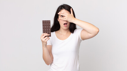 pretty woman looking shocked, scared or terrified, covering face with hand and holding a chocolate...