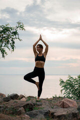 Woman in black sport suit is doing yoga pose (asana), work out outside near beautiful river and beautiful landscape with wild flowers and rocks