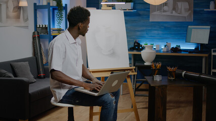 Man of african american ethnicity working on vase drawing using laptop computer in art studio. Black young artist doing research online for masterpiece inspiration and creativity © DC Studio