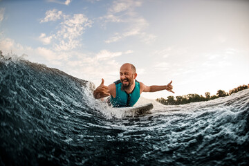 cheerful male wakesurfer lying on board rides down wave and show hands gestures and winks
