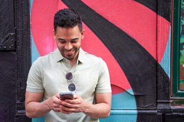 Cheerful young latin guy texting, checking social networks, chatting on his smart phone on colorful...