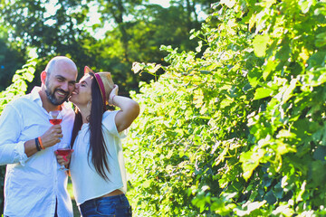 Naklejka premium Portrait of a smiling happy couple kissing in a Vineyard toasting wine. Beautiful brunette woman and bearded muscular man spending time together