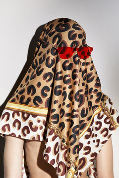 Staged photo of slim lady with beige scarf thrown over her head and fixed with red sunglasses. Stylish head scarf is made of silk fabric with leopard pattern. 