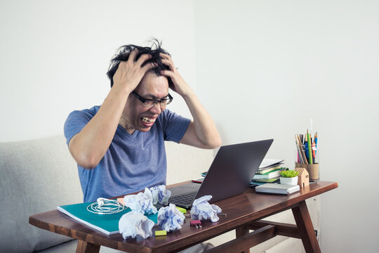 Crazy shouting man with messy hair, wide open mouth and holding hands on head sitting at home table using laptop. Work from home