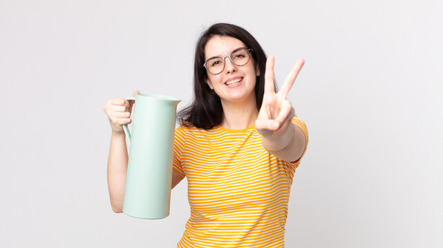 pretty woman smiling and looking happy, gesturing victory or peace and holding a coffee thermos