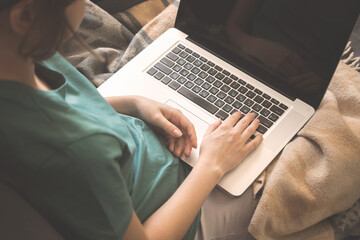 Young girl using laptop sitting on sofa with warm plaid indoors. Hands on keyboard, black screen