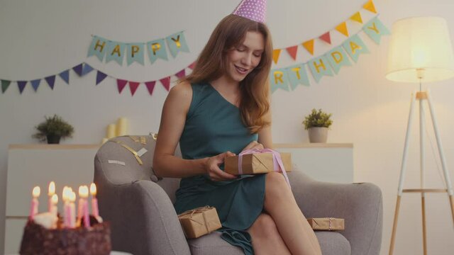Birthday party celebration. Young happy woman unpacking gift box, sitting on armchair in decorated room