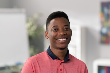 Portrait of young boy with dark eyes, hair, black skin, beautiful wide white smile, blurred background, man is wearing salmon collar shirt, student in class is looking at camera