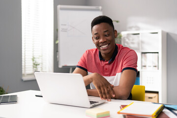 Fototapeta premium Trainee in office sits at desk in front of laptop monitor in background whiteboard, young dark-skinned man talks online with clients, co-workers, uses webcam, video call, smiling, cheerful