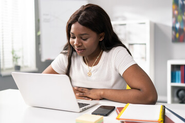 Modest, pretty, dark-skinned girl sits in room at desk, spends afternoon chatting with friends, surfing internet, next to phone, notebooks, women smiles softly at laptop screen, wainting for messages