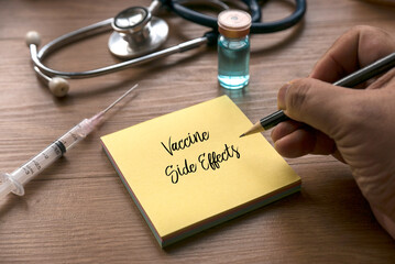 A bottle of vaccine, syringe, stethoscope and a hand writing on yellow memo note written with...