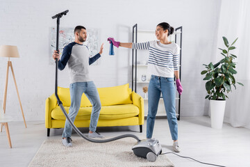 African american woman holding detergent near boyfriend with vacuum cleaner