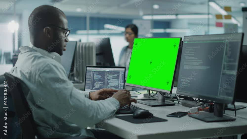 Fototapete Multi-Ethnic Office: Black IT Programmer Working on Computer with Green Screen Chroma Key Display. Male Software Engineer Developing App, Program, Video Game. Terminal with Code Language. Static Shot