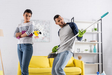 Man playing on mop near offended african american girlfriend in rubber gloves