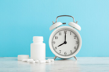 White alarm clock and plastic bottle with pills on table at light blue wall background. Pastel...