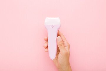 Young adult woman hand holding electric epilator on light pink table background. Pastel color. Closeup. Female product for smooth body skin. Top down view.