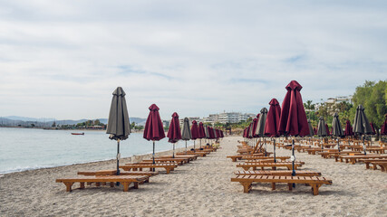 Empty wooden deck beds and folded umbrellas on sand near Aegean Sea. Lounge chill zone on city...