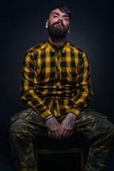 Tattooed man with a beard wearing checkered shirt is posing seated on a chair in a studio