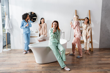 cheerful interracial women holding cosmetic products in bathroom during slumber party