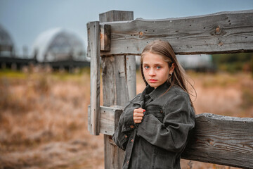A sad little girl wrapped in an oversized denim jacket standing near a wooden fence. The concept of...