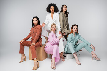 full length of interracial models in stylish suits sitting while posing on grey