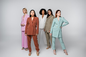 full length of interracial models in stylish pastel suits posing on grey