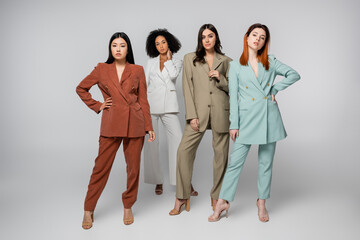 full length of multiethnic models in pastel color suits posing on grey