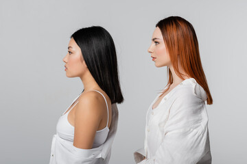 side view of brunette asian woman and redhead model posing isolated on grey