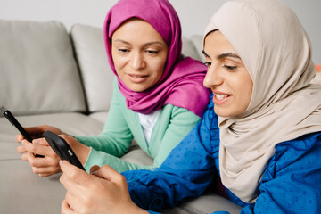 Fototapeta premium Two women in headscarf using mobile phones while resting on sofa