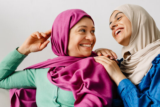 Two women in headscarf hugging and laughing while sitting on sofa