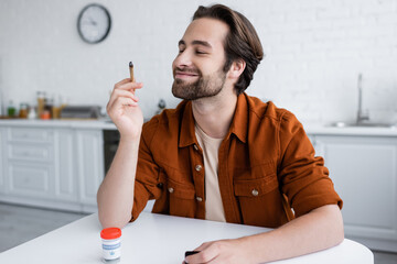Smiling man looking at joint of medical cannabis near jar on table