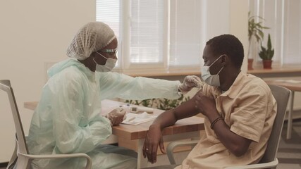 Medium shot of female medical worker in protective mask and clothing doing Covid-19 vaccine shot in arm of young African American man