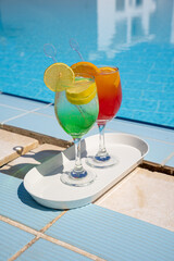 two wine glasses with red cocktail or juice with bendie and piece of orange on edge, water and swimming pool behind
