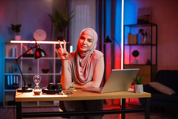 Night lighted home remote office. Front view of gorgeous smiling arabic businesswoman in hijab, sitting at the desk with laptop and gesturing ok sign into camera