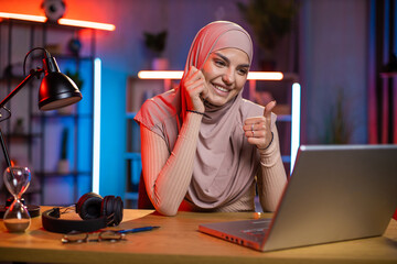 Positive arabian woman sitting at desk during evening time and chatting online with friends via video call. Young female in hijab using modern laptop for remote work at home.