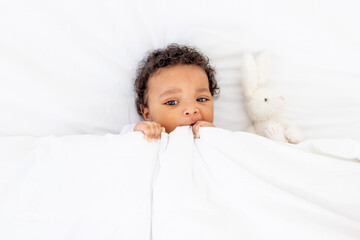 Smiling African-American little baby under a blanket with a toy on a white bed for sleeping