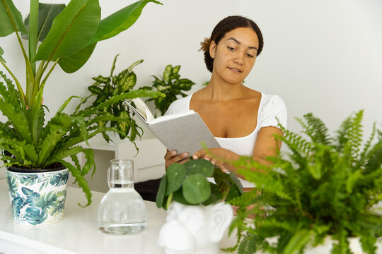 Thoughtful ethnic woman with notebook between assorted plants at home