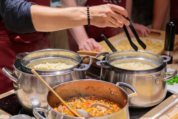 The chef prepares pasta in large metal pans. Male hands take out the pasta from the pan with tongs. Close-up.