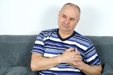 old bald man sitting on the couch, clasped his head in his hands, suffering on his face, concept of heart problems, coronary artery disease, heart attack