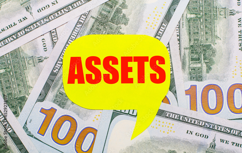 Poster against the background of dollars scattered on the table is a yellow curly card with the text assets