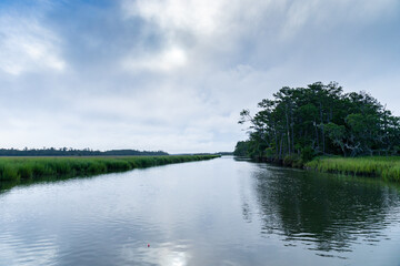 Partially overcast early morning light on a waterway through a grassy green salt marsh, distant boats and trees, horizontal aspect