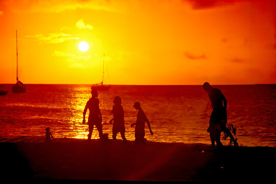 Plage des Caraïbes dans une ambiance jaune orange du coucher de soleil, des bateaux en toile de fond et des personnes sur le sable.
