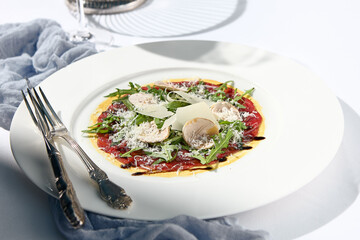 Beef carpaccio with mushrooms and parmesan cheese. Fancy dinning with beef carpaccio on white table with simple contemporary decor. Sunlight and harsh shadow still life. Beef carpaccio appetizer dish.