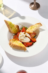 Traditional Italian salad with Burrata cheese and tomatoes. Fancy caprese with burrata cheese, cherry tomatoes, pesto and balsamic sauce. Summer dish with mozzarella cheese on white table with shadow.