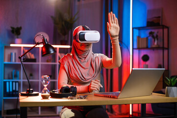Fototapeta na wymiar Young arabian woman sitting at home office and using VR headset and laptop for work during evening time. Concept of people, technologies and freelance.
