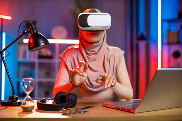 Young woman wearing VR goggles while sitting at desk with wireless laptop. Pretty lady in hijab touching with fingers virtual screen. Modern technologies for work at home.