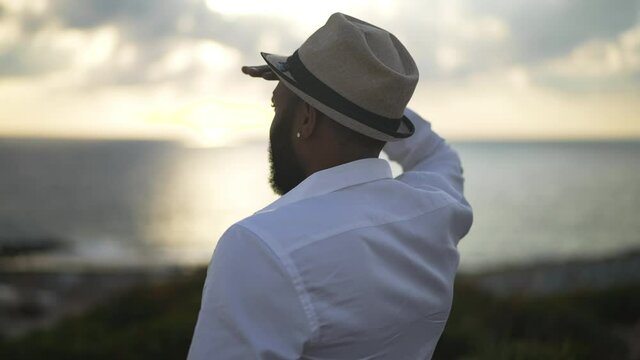 Male African American tourist in hat and white shirt looking at sunrays at sunset standing on Mediterranean sea coast. Relaxed happy LGBT gay man admiring beauty of nature outdoors at resort