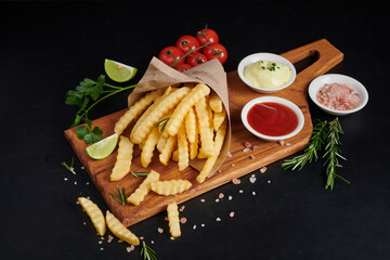 Homemade baked potato fries with mayonnaise, tomato sauce and rosemary on wooden board. tasty french fries on cutting board, in brown paper bag on black stone table background, unhealthy food.