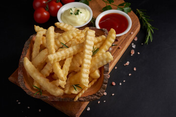 Homemade baked potato fries with mayonnaise, tomato sauce and rosemary on wooden board. tasty french fries on cutting board, in brown paper bag on black stone table background, unhealthy food.